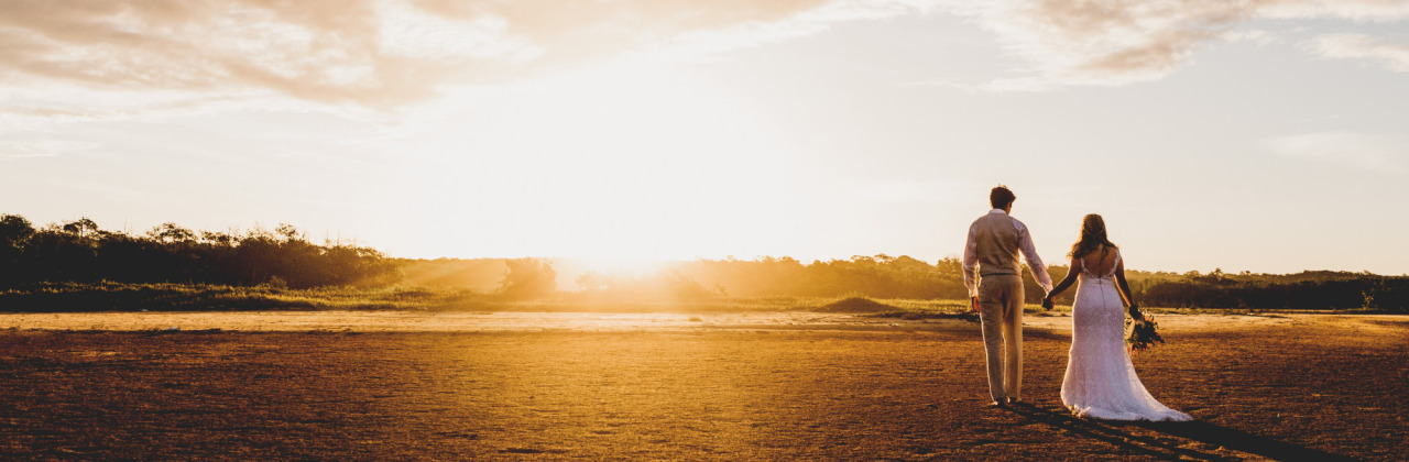 A man and woman holding hands stood in a field looking at the sun and sky