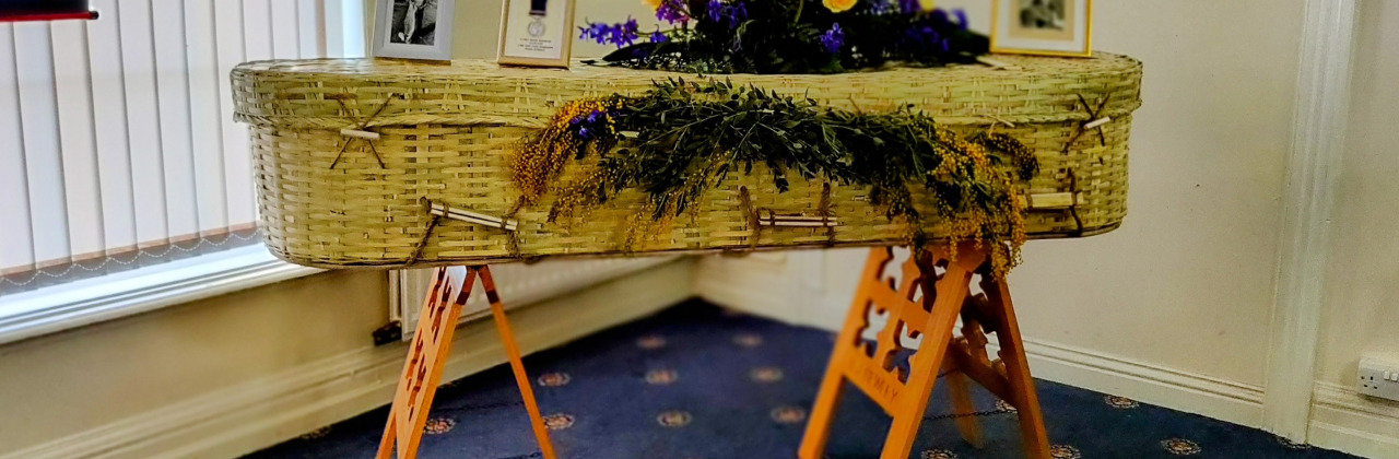 A wicker coffin with flowers and photos on a stand