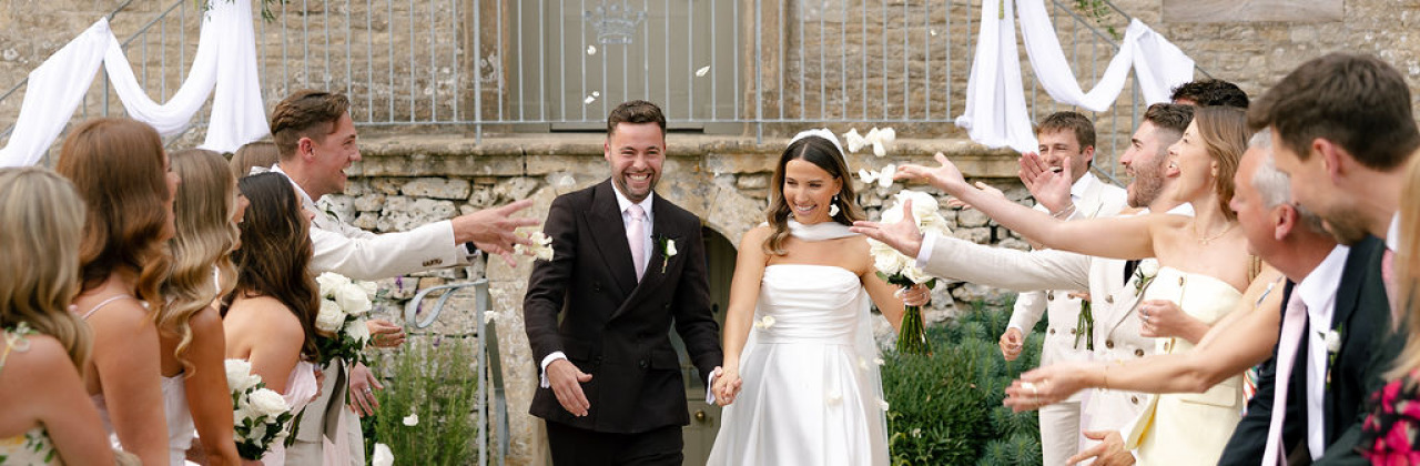 A photo of a couple making their way down a confetti corridor in front of the facade of Ivy Lodge in Cirencester.