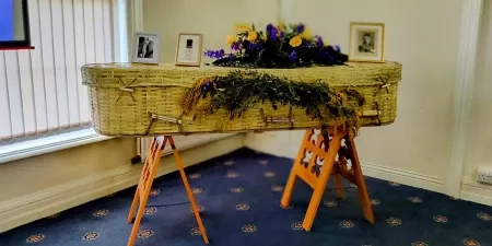 A wicker coffin with flowers and photos on a stand