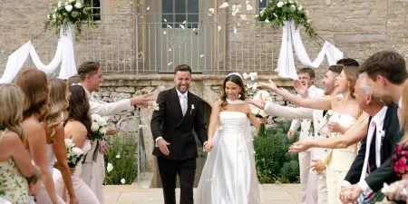 A photo of a couple making their way down a confetti corridor in front of the facade of Ivy Lodge in Cirencester.