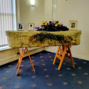 A wicker coffin with flowers and photos on a stand