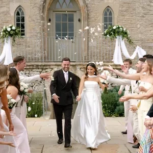 A photo of a couple making their way down a confetti corridor in front of the facade of Ivy Lodge in Cirencester.